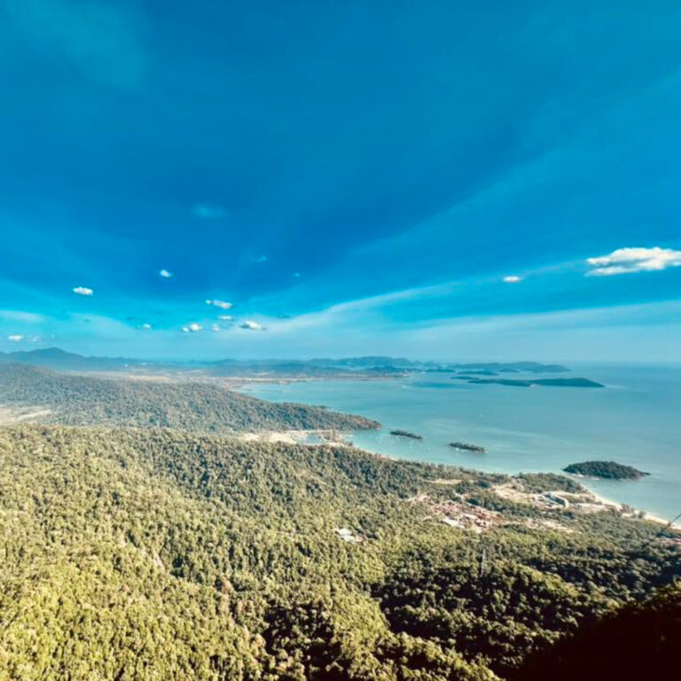 Langkawi coastline view from above, lush green hills meet the turquoise sea under a bright blue sky.
