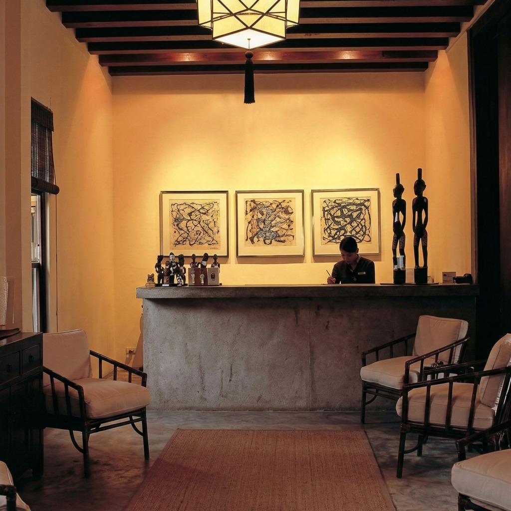 Hotel reception at Ratchamankha, featuring artwork, wooden chairs, and a dark-haired receptionist.
